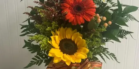 A tall mixed floral bouquet with sunflowers, a red gerbera daisy, and white blooms in a glass vase.