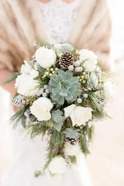 A close-up of a wedding bouquet held by a bride, featuring white roses, green succulents, and pinecones.