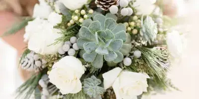 A close-up of a wedding bouquet held by a bride, featuring white roses, green succulents, and pinecones.
