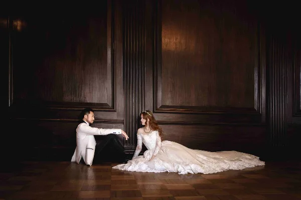 A bride in a white lace gown and a groom in a white tuxedo jacket sitting on a wooden floor against a dark paneled wall.