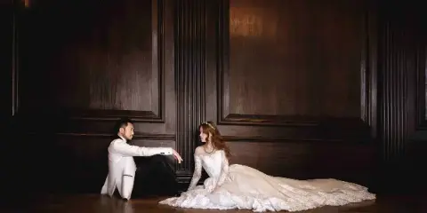 A bride in a white lace gown and a groom in a white tuxedo jacket sitting on a wooden floor against a dark paneled wall.