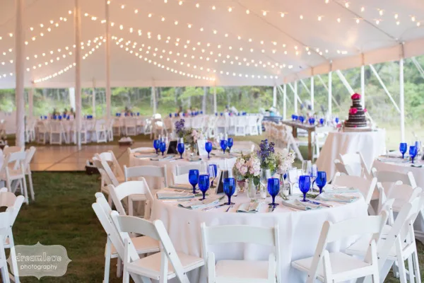 Interior of a white event tent set up with round tables, white chairs, blue glassware, and string lighting.