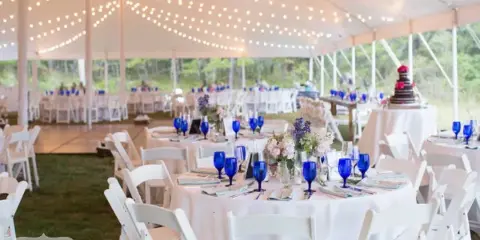 Interior of a white event tent set up with round tables, white chairs, blue glassware, and string lighting.
