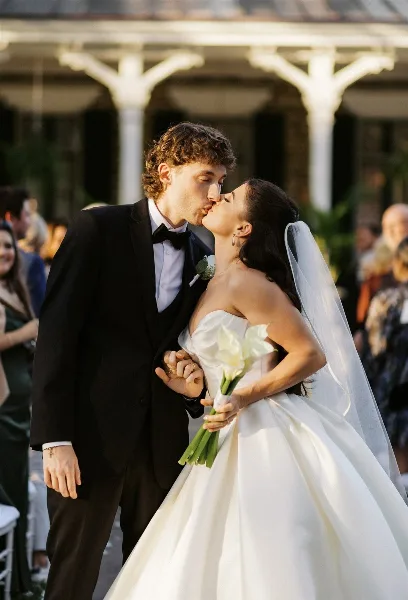 A bride and groom share a kiss during their wedding ceremony, with the bride holding a bouquet of white calla lilies.