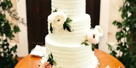 A three-tiered white wedding cake with horizontal ridges and floral decorations on a wooden table.