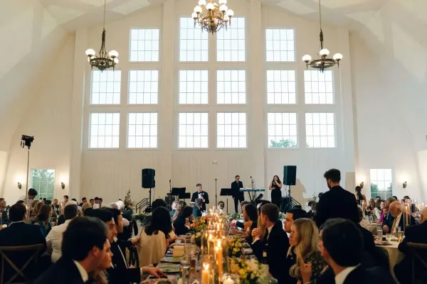 Interior view of a wedding reception in a white barn with high ceilings, chandeliers, and guests seated at long tables.