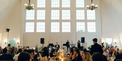 Interior view of a wedding reception in a white barn with high ceilings, chandeliers, and guests seated at long tables.