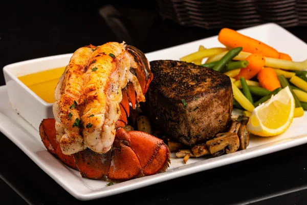 A plated meal featuring a lobster tail, a thick-cut steak, and a side of mixed vegetables on a white rectangular plate.