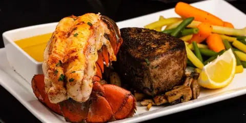 A plated meal featuring a lobster tail, a thick-cut steak, and a side of mixed vegetables on a white rectangular plate.