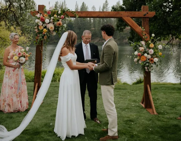 A bride and groom exchanging rings under a wooden arch decorated with flowers during an outdoor wedding ceremony.