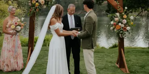 A bride and groom exchanging rings under a wooden arch decorated with flowers during an outdoor wedding ceremony.