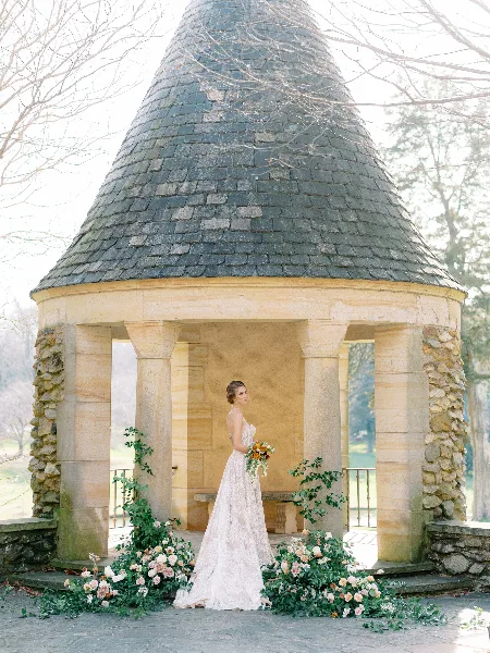A bride in a white lace wedding dress and styled updo stands in a stone gazebo holding a bouquet.