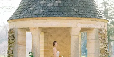 A bride in a white lace wedding dress and styled updo stands in a stone gazebo holding a bouquet.