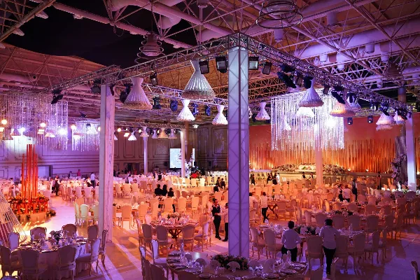 A wide shot of a large event hall featuring professional lighting trusses, hanging crystal chandeliers, and numerous round tables set for a formal gathering.