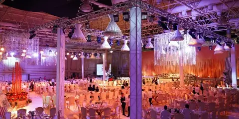 A wide shot of a large event hall featuring professional lighting trusses, hanging crystal chandeliers, and numerous round tables set for a formal gathering.