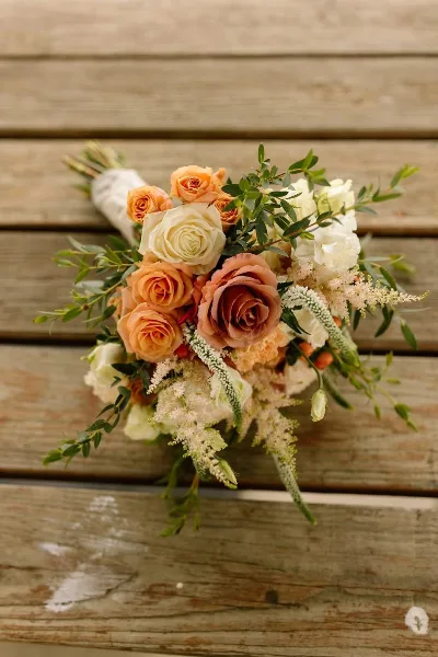 A wedding bouquet with peach and white roses, greenery, and filler flowers resting on a wooden surface.