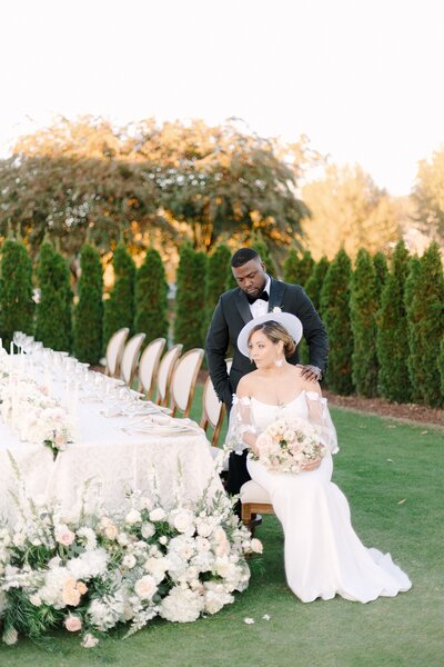 A bride and groom posing together at an outdoor wedding reception with a long floral table setting.