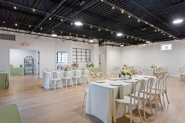 Interior view of a wedding venue with white tables, chairs, and floral centerpieces under a dark ceiling with string lights.