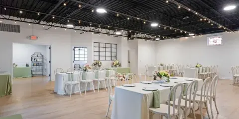 Interior view of a wedding venue with white tables, chairs, and floral centerpieces under a dark ceiling with string lights.