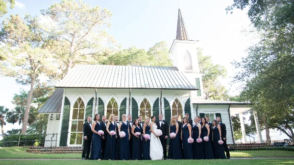 A wedding party posing in front of a white church with a steeple.