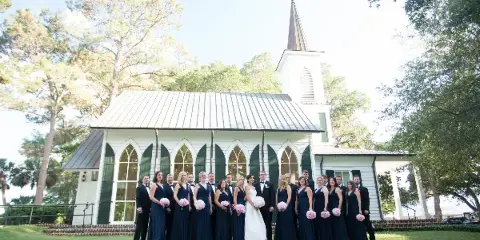 A wedding party posing in front of a white church with a steeple.