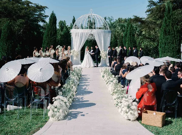 A wide shot of an outdoor wedding ceremony with a bride and groom at a white domed altar, surrounded by guests holding white parasols and floral arrangements.
