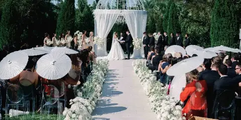 A wide shot of an outdoor wedding ceremony with a bride and groom at a white domed altar, surrounded by guests holding white parasols and floral arrangements.