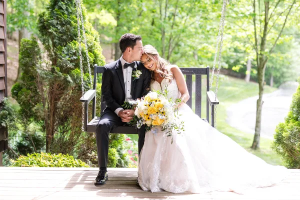 A bride and groom sitting on a wooden swing outdoors, with the groom kissing the bride's forehead.