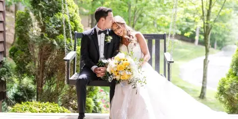 A bride and groom sitting on a wooden swing outdoors, with the groom kissing the bride's forehead.