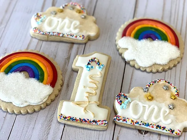 A collection of custom-decorated sugar cookies featuring rainbow designs and the number one, arranged on a light wooden surface.