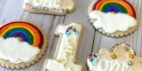 A collection of custom-decorated sugar cookies featuring rainbow designs and the number one, arranged on a light wooden surface.