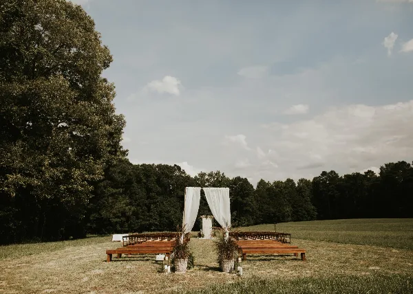 Outdoor wedding ceremony setup with a white draped arch, rows of wooden benches, and potted plants in a grassy field.