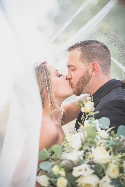 A bride and groom sharing a kiss under a veil, with a bouquet of white flowers in the foreground.