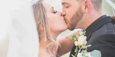 A bride and groom sharing a kiss under a veil, with a bouquet of white flowers in the foreground.