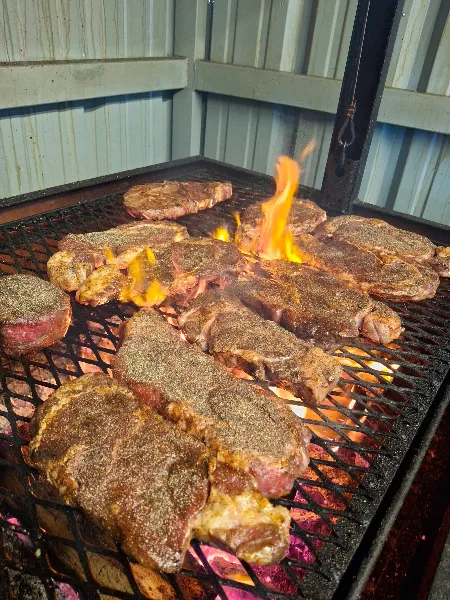 Several seasoned steaks cooking on a grill grate over glowing coals and open flames.