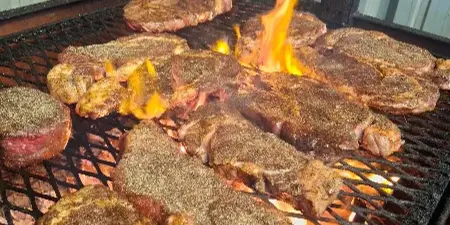 Several seasoned steaks cooking on a grill grate over glowing coals and open flames.