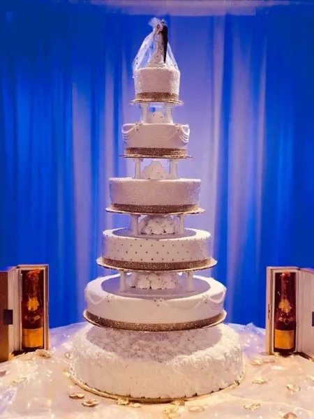 A tall, multi-tiered white wedding cake with gold accents and a bride and groom topper, displayed against a blue curtain backdrop.