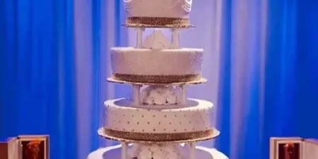 A tall, multi-tiered white wedding cake with gold accents and a bride and groom topper, displayed against a blue curtain backdrop.