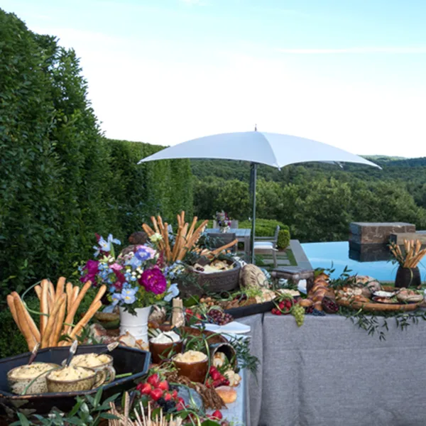 An outdoor catering table set with breadsticks, dips, and floral centerpieces under a white umbrella.
