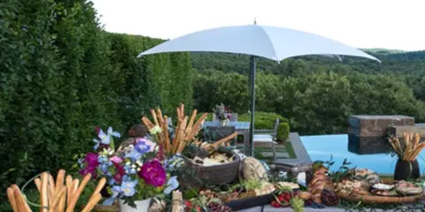 An outdoor catering table set with breadsticks, dips, and floral centerpieces under a white umbrella.
