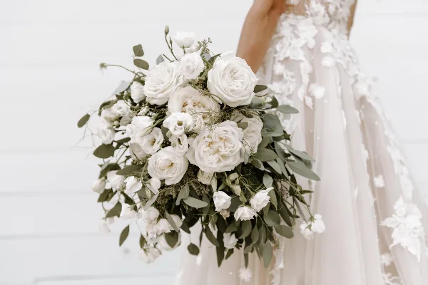 A close-up photograph of a bride holding a cascading white bouquet featuring roses, eustoma, and eucalyptus foliage.