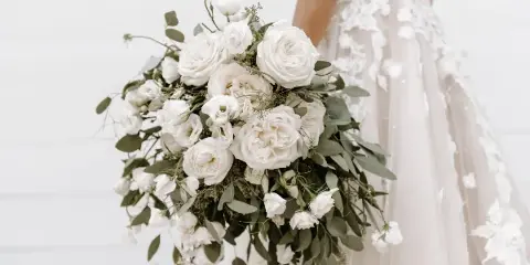 A close-up photograph of a bride holding a cascading white bouquet featuring roses, eustoma, and eucalyptus foliage.