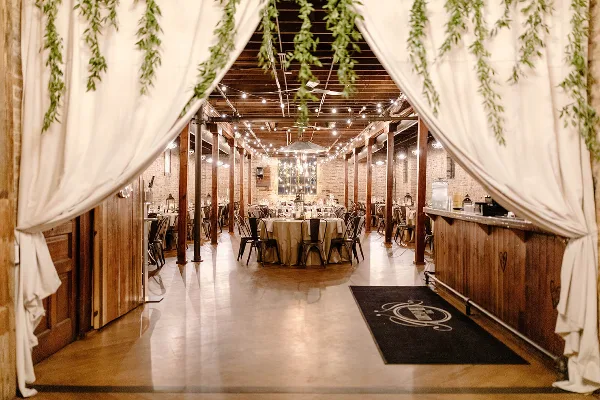 Interior of an industrial-chic wedding venue with exposed brick walls, wood beams, string lights, and a reception table setup.