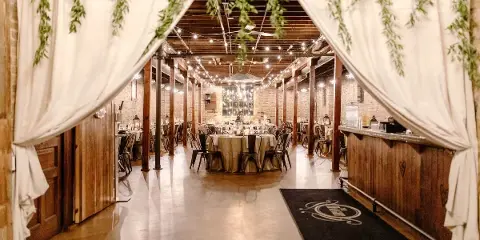 Interior of an industrial-chic wedding venue with exposed brick walls, wood beams, string lights, and a reception table setup.
