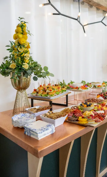 A catering buffet table featuring a tall lemon and eucalyptus centerpiece, skewered appetizers, and a charcuterie board.