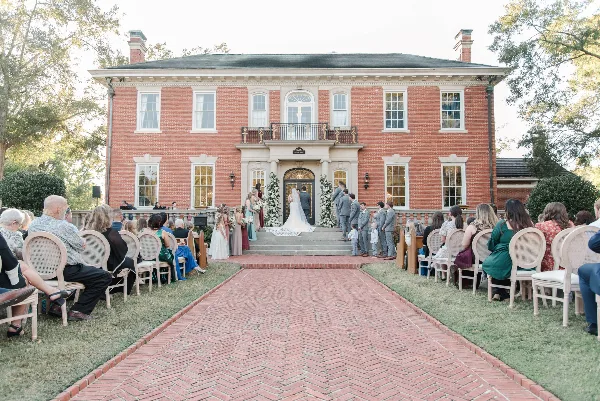 A wide shot of a wedding ceremony taking place on the brick front steps of a large red brick historic house, with guests seated on either side of a brick aisle.