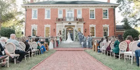 A wide shot of a wedding ceremony taking place on the brick front steps of a large red brick historic house, with guests seated on either side of a brick aisle.