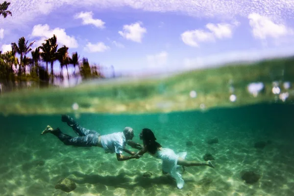A couple swimming underwater in a tropical location with palm trees visible above the water surface.
