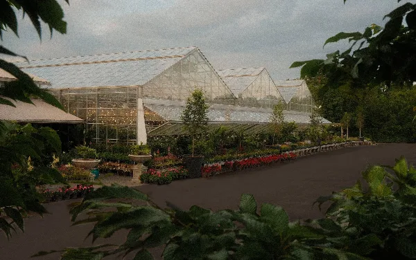Glass greenhouse structure at a plant nursery with rows of potted plants and flowers along a paved walkway.
