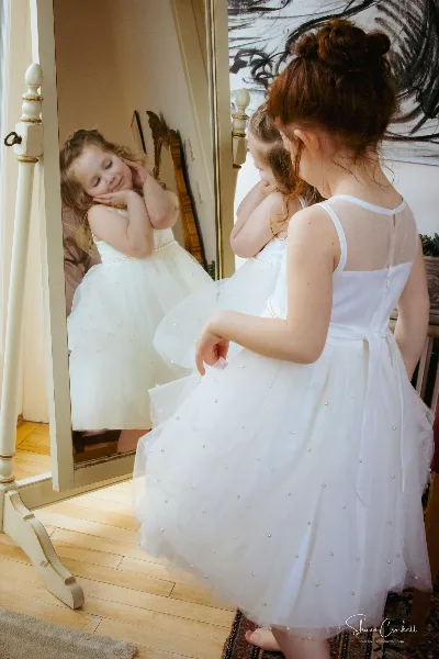 A young girl in a white dress looking at her reflection in a mirror, photographed by Shaun Crockett.